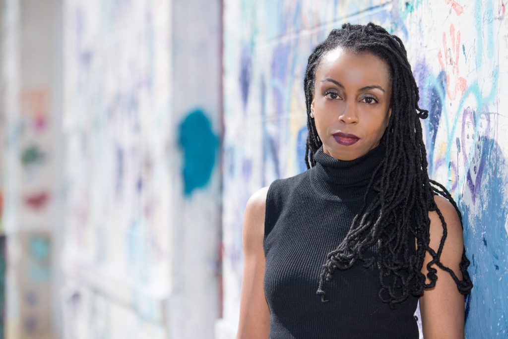 A headshot of Morowa Yejidé standing in front of a painted wall wearing a black, high-neck top.