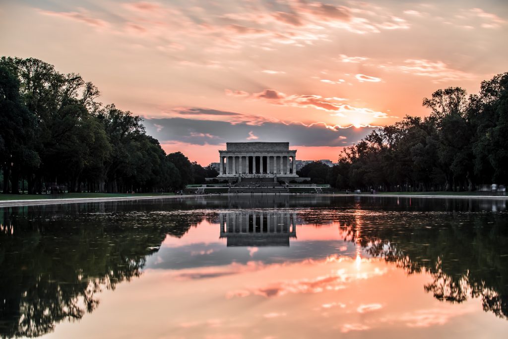 A picture of the Lincoln Memorial in Washington, D.C. over the famous reflecting pool, and its reflection is shown in the water. The sky is pink, and the sun peeks out from behind a cloud.