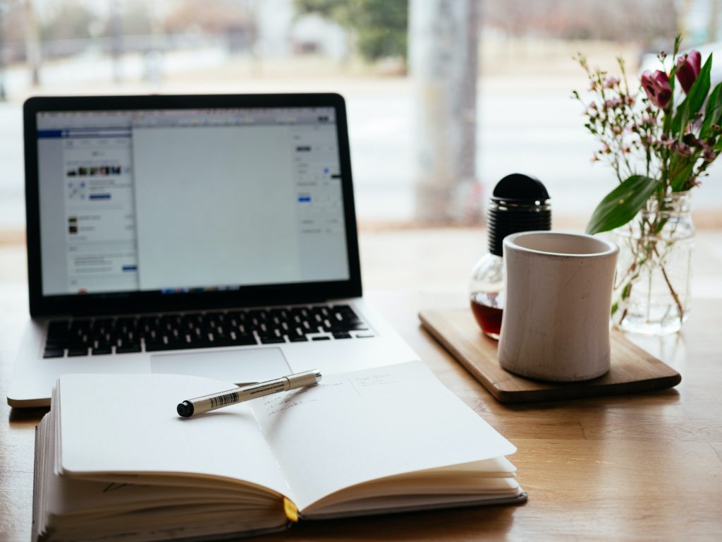 A photo of a desk at a coffee shop, and on it are a notebook and pen, a laptop, a coffee mug, and a vase of flowers.