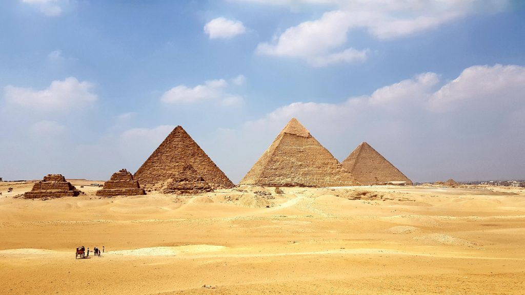 A photo of three large pyramids and three small pyramids in an Egyptian desert. The ground is tan and sandy and the sky is bright blue with scattered clouds.