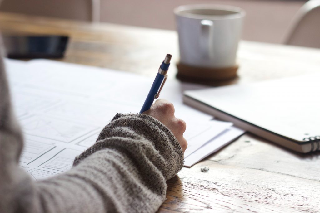 A person writing on paper with a coffee mug in the background.