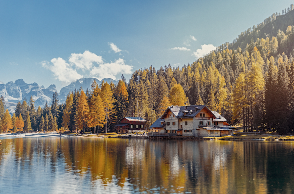 A mountainous area with a variety of green and yellow pine trees reflecting over a clear lake. There's a log cabin on the lake shore.