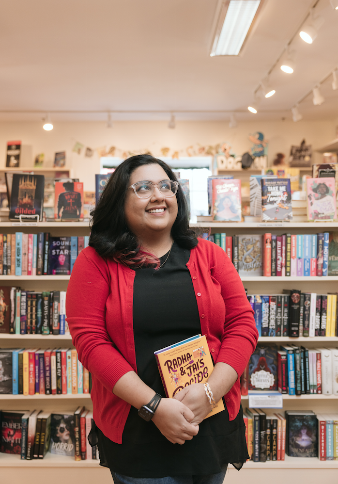 Photo of Nisha Sharma smiling and holding her book while standing in front of book stacks.