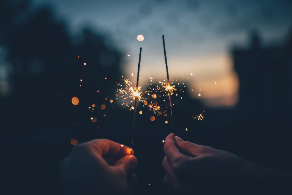 Two hands holding lit sparklers as the sun sets.