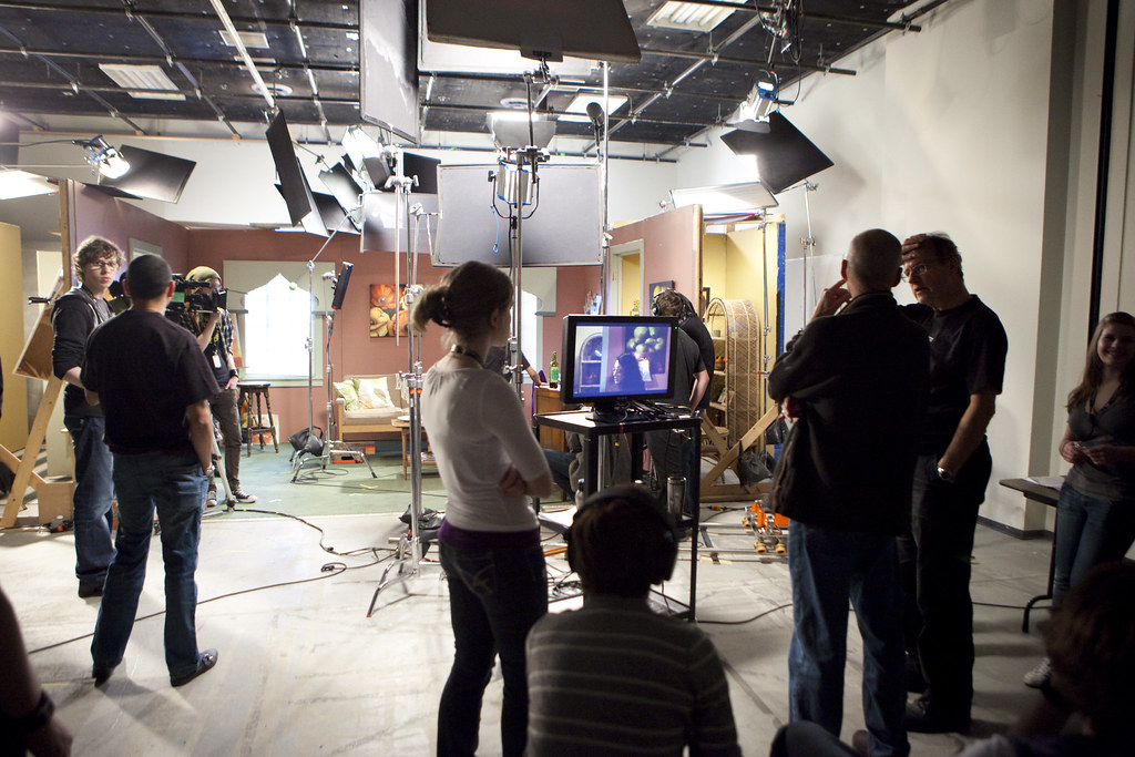 This is a photo of a group of students in a classroom film studio. A woman is working the camera while another group of students stand nearby.