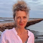 This picture shows Alice O'Neill, a new faculty member at Wilkes University. She is wearing a white linen shirt and she is standing on what looks like a beach at low tide. Her blonde, curly hair is piled in an updo on the top of her head. She is smiling.